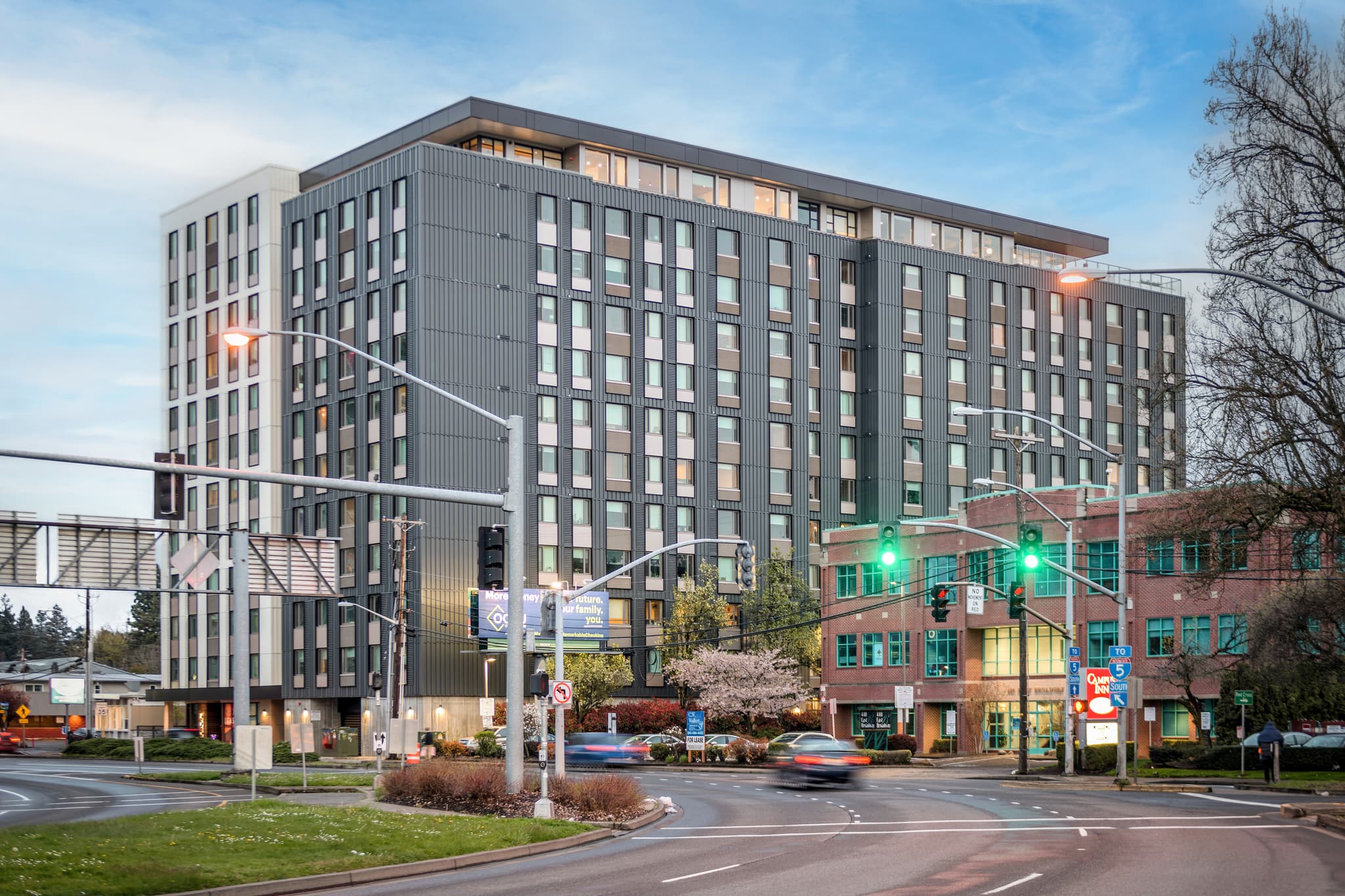 Union on Broadway is a modern multi-story building with large windows, positioned at a busy intersection with streetlights, traffic signals, crosswalks, and sidewalks. Trees and greenery surround the area beneath a partly cloudy sky.