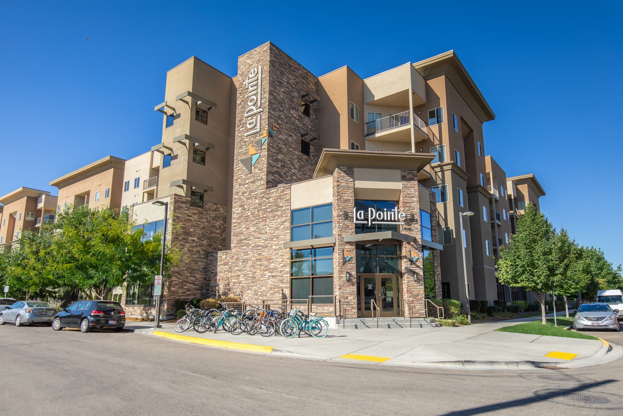 A modern apartment building with a La Pointe sign offers off-campus student housing in Boise. Featuring a stone and tan exterior, balconies, and a bike rack outside, cars are parked along the street under a clear blue sky.