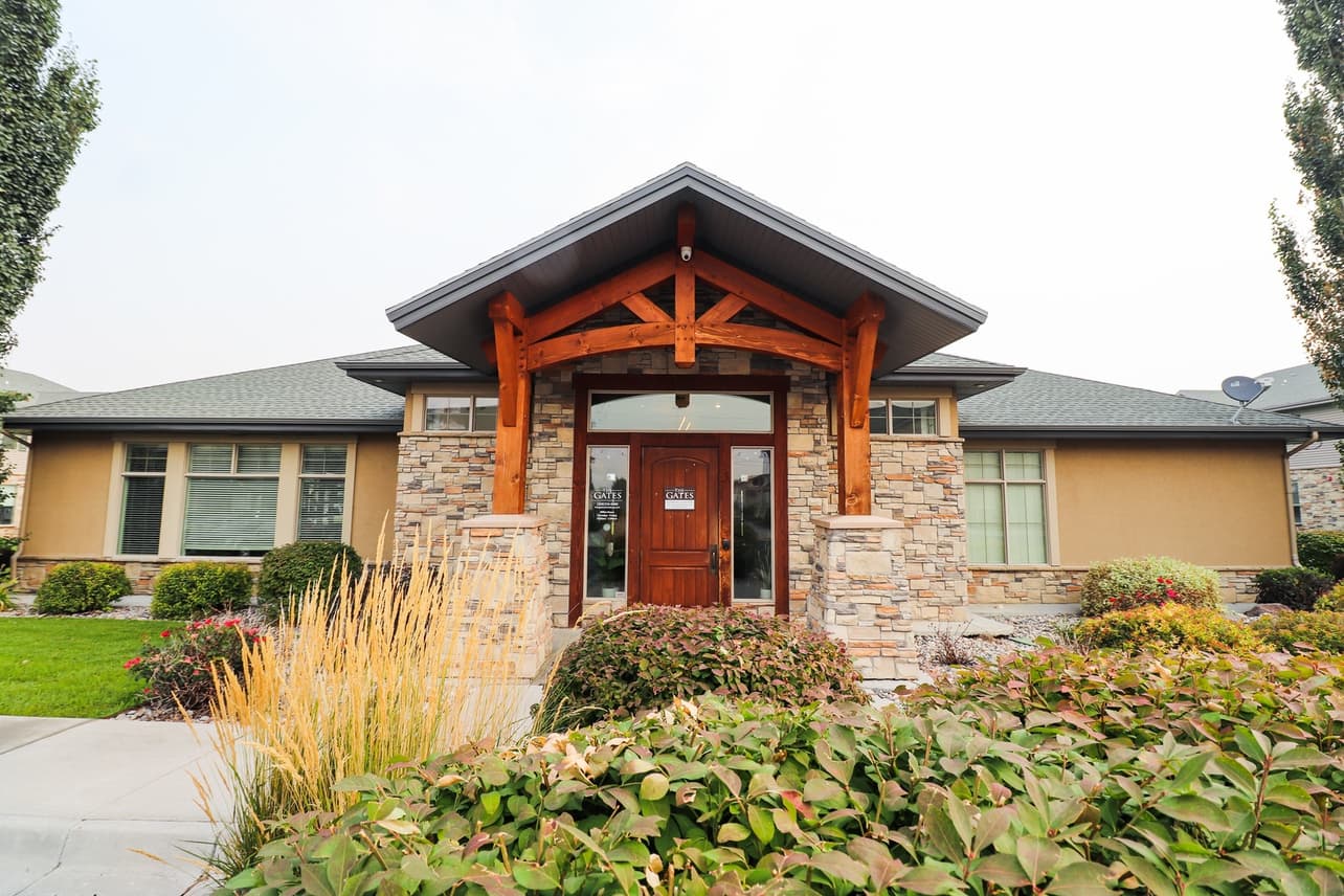 Single-story house at The Gates at Rexburg with stone and tan siding, a wooden front door, exposed wooden beams, large windows, and a landscaped front yard featuring bushes, grasses, and a concrete path.