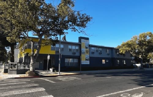 A two-story apartment building with gray and yellow accents sits on a street corner, surrounded by trees under a clear blue sky. A large vertical sign reading "Heritage 2831" is displayed on the building's side.