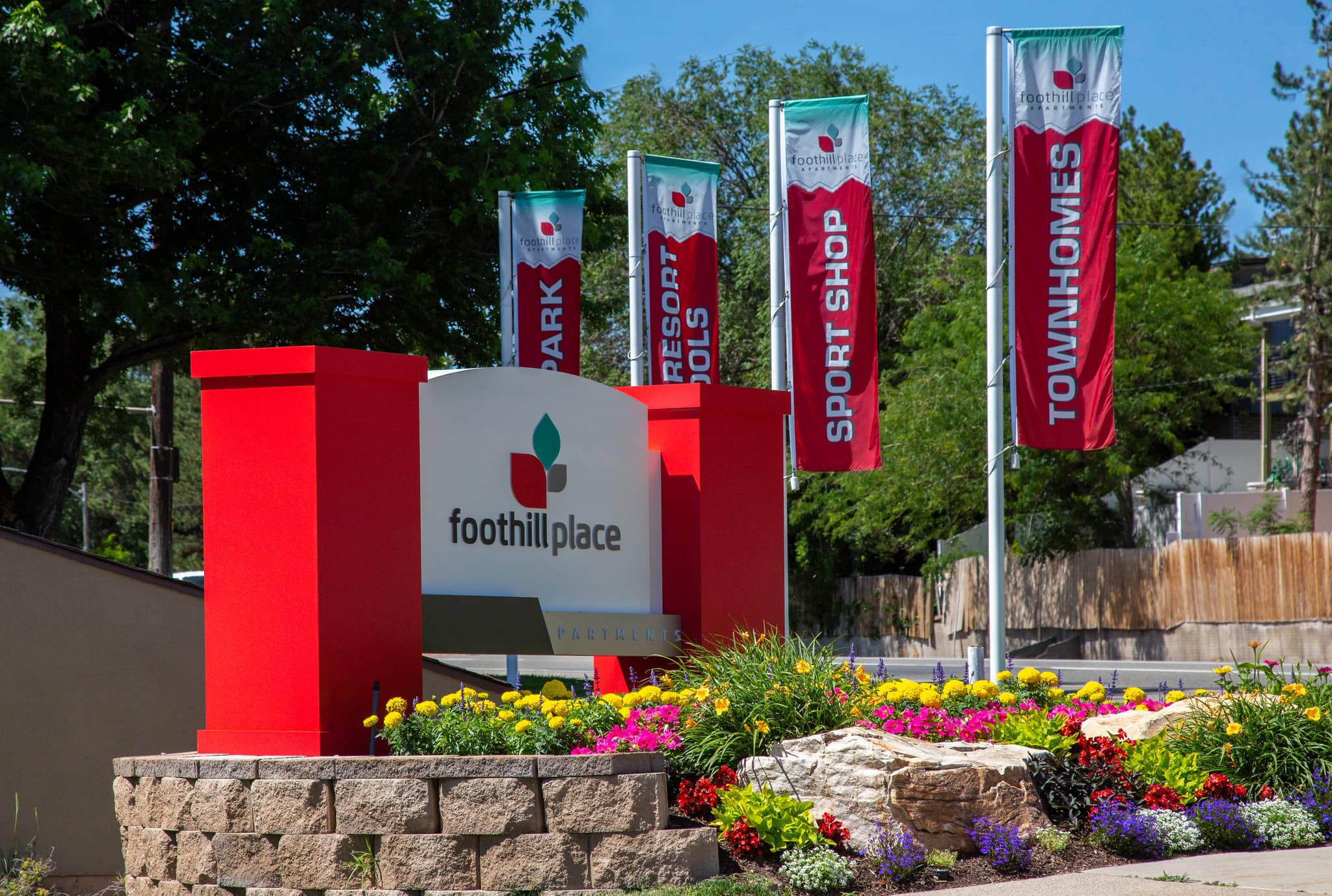 Foothill Place Salt Lake City entrance sign with orange pillars, surrounded by colorful flower landscaping. Four banners in the background advertise Park, Resort Pools, Sport Shop, and Townhomes. Trees and blue sky complete the scene.