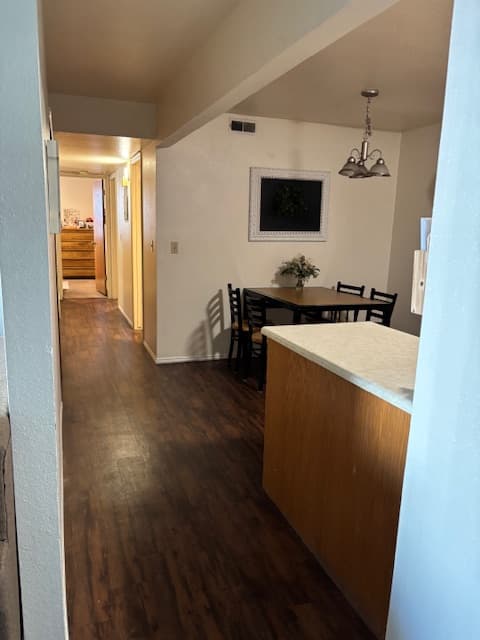 A hallway with wood flooring leads to a bedroom at Carriage Cove Provo. To the right, there’s a small dining table with four chairs under a hanging light fixture. A kitchen counter with a light-colored countertop is in the foreground.