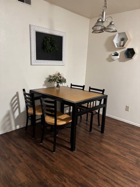 A dining area at Carriage Cove Provo features a wooden table and four chairs on a dark wood floor. A vase with flowers sits on the table, while the walls display a framed wreath and geometric shelves with small plants.