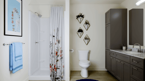 A modern bathroom at Ivory University House Salt Lake City features a shower with a floral curtain, white toilet, dark gray cabinets, marble countertop, circular blue rug, and three geometric plant holders. Towels and art complete the stylish space.
