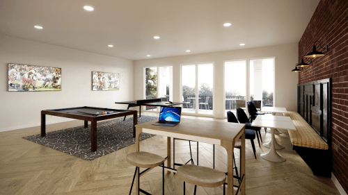 A modern game room at Ivory University House Salt Lake City features large windows, a pool table, a laptop-ready table, bench seating against a brick wall, wall art, and tree views. The bright space is enhanced by recessed lighting.