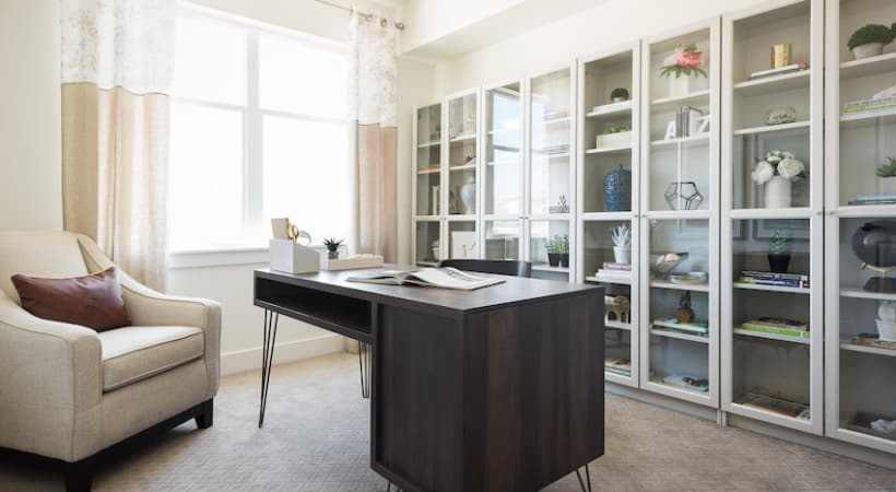 A modern home office with a dark wood desk, beige armchair, and glass-fronted bookshelves filled with books and plants captures the tranquil vibe of Arboretum at Barber Station Boise as sunlight streams in from a large window with cream curtains.