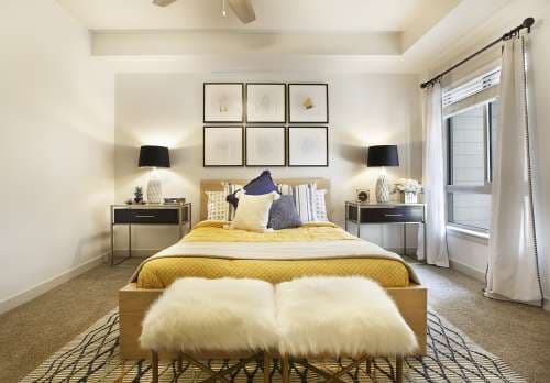 A modern bedroom with a wooden bed, yellow bedding, pillows, benches with white fur covers, bedside tables with black lamps, framed art above the bed, and a large window with sheer curtains—perfect for a stylish retreat on Liberty Blvd, Salt Lake City.