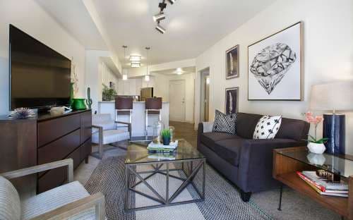 Modern living room with a dark sofa, glass coffee table, large TV, and diamond-themed wall art. Neutral decor includes a rug, lamps, plants, and a view into the kitchen and dining area—perfect for stylish living on Liberty Blvd in Salt Lake City.