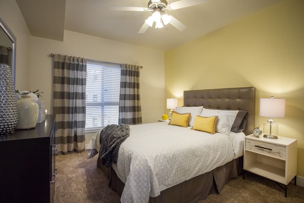 A cozy bedroom at Liberty Gateway Salt Lake City features a large bed with a gray tufted headboard, white and yellow pillows, two nightstands with lamps, striped curtains on the window, and neutral decor under a ceiling fan.