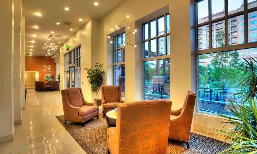 Modern hotel lobby at Liberty Gateway Salt Lake City features large windows, orange patterned armchairs around a small table, indoor plants, and bright ceiling lights. Sunlight streams in with views of tall buildings outside.