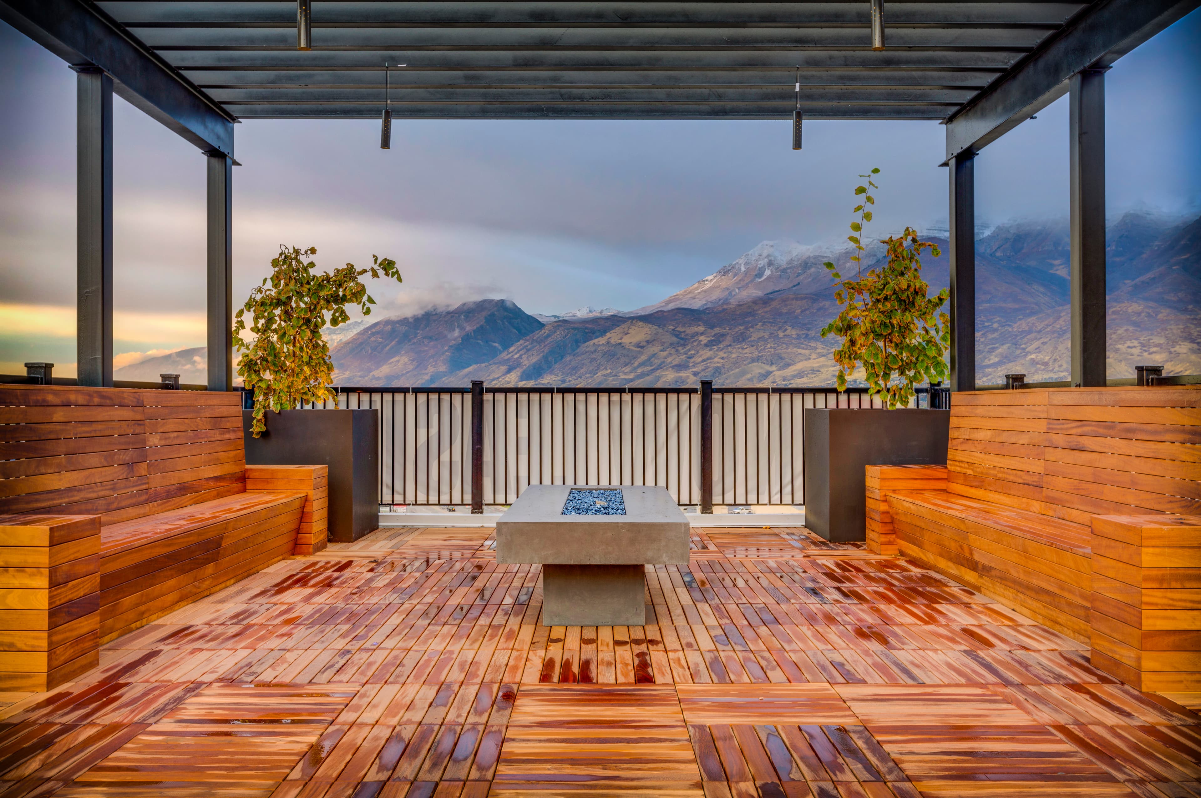 A modern outdoor patio at Midtown 360 Orem features wooden benches and a central fire pit table, overlooking snow-capped mountains beneath cloudy skies. Two potted plants sit in the far corners under a stylish pergola roof.