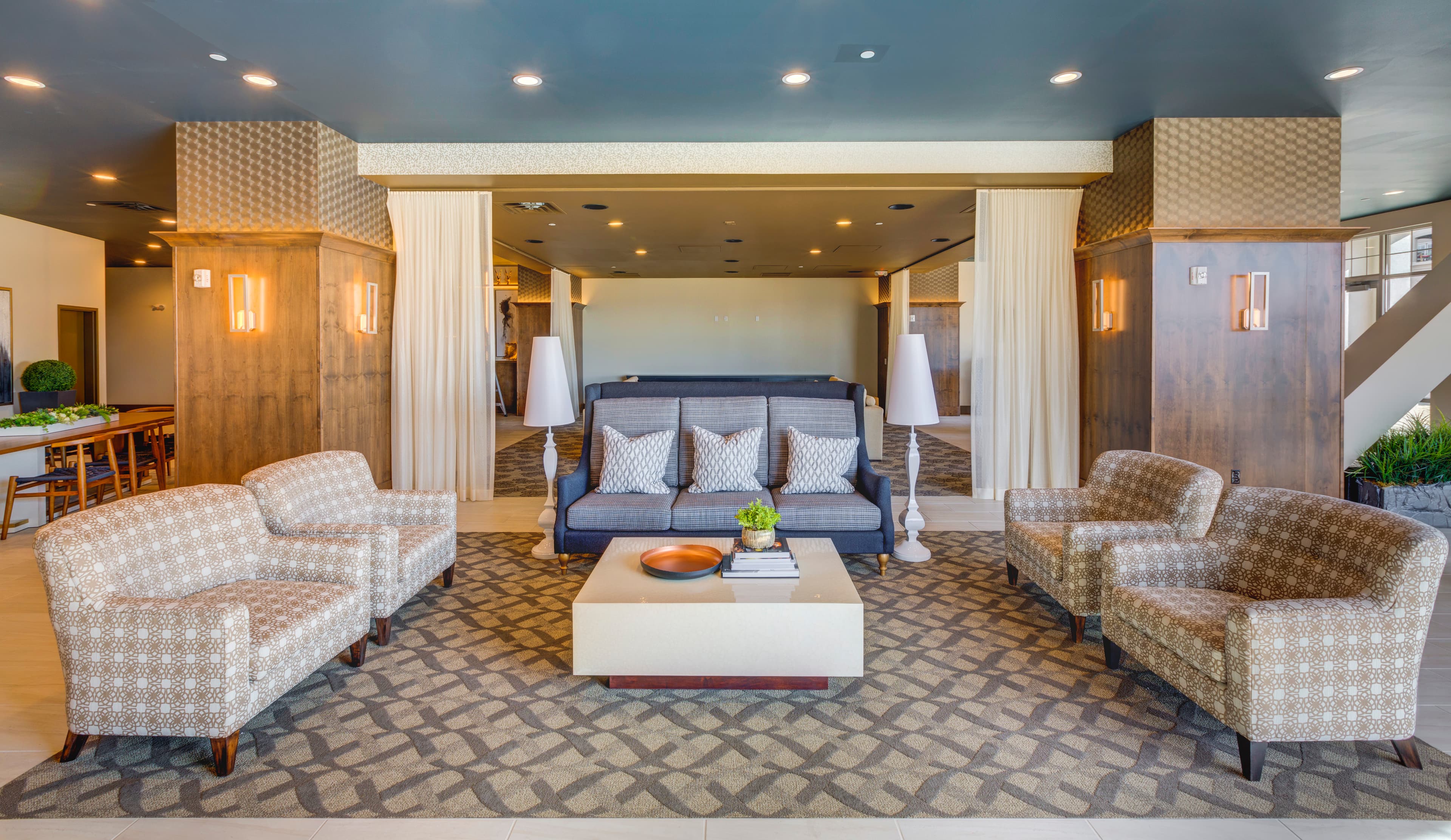 The Midtown 360 Orem hotel lobby features patterned armchairs, a gray sofa, a geometric rug, and a rectangular coffee table with books and decor. Warm lighting, decorative pillars, and neutral colors create an inviting atmosphere.