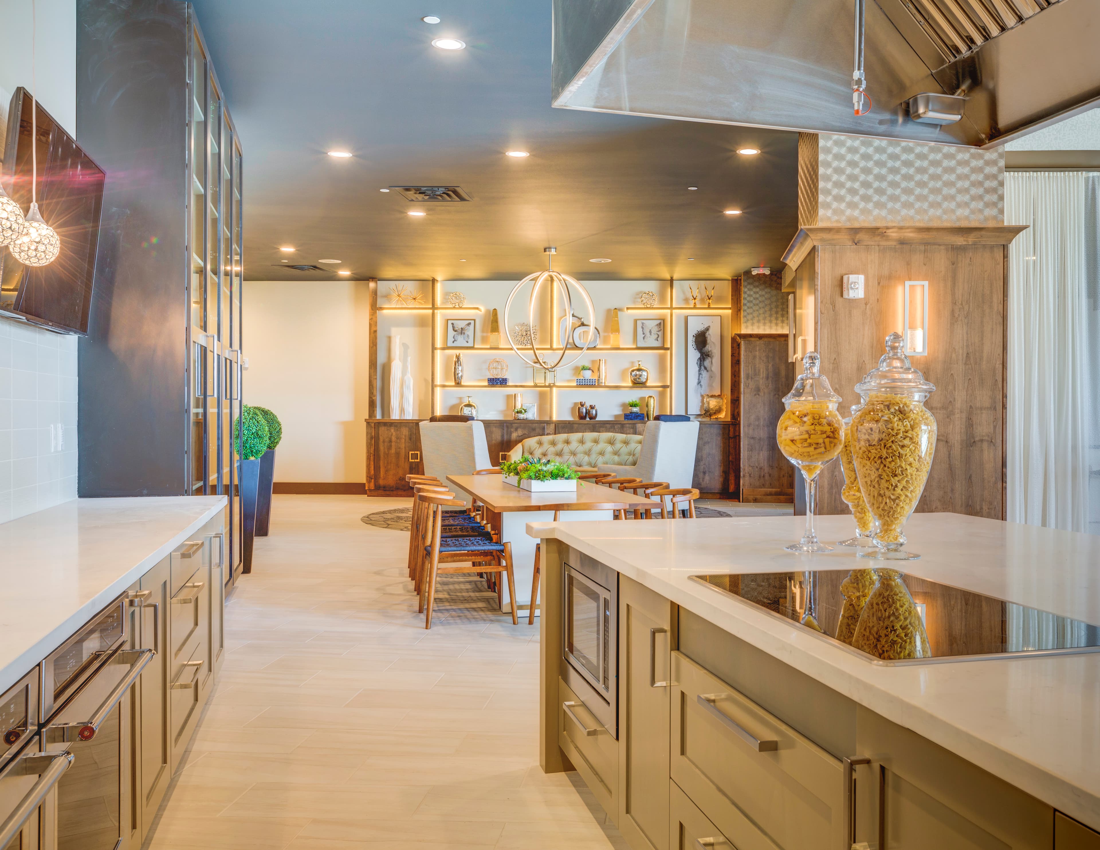 Modern kitchen and dining area at Midtown 360 Orem features light wood floors, stainless steel appliances, glass pasta jars on the island, and contemporary lighting. A dining table with chairs and a green cushioned bench sit in the background.