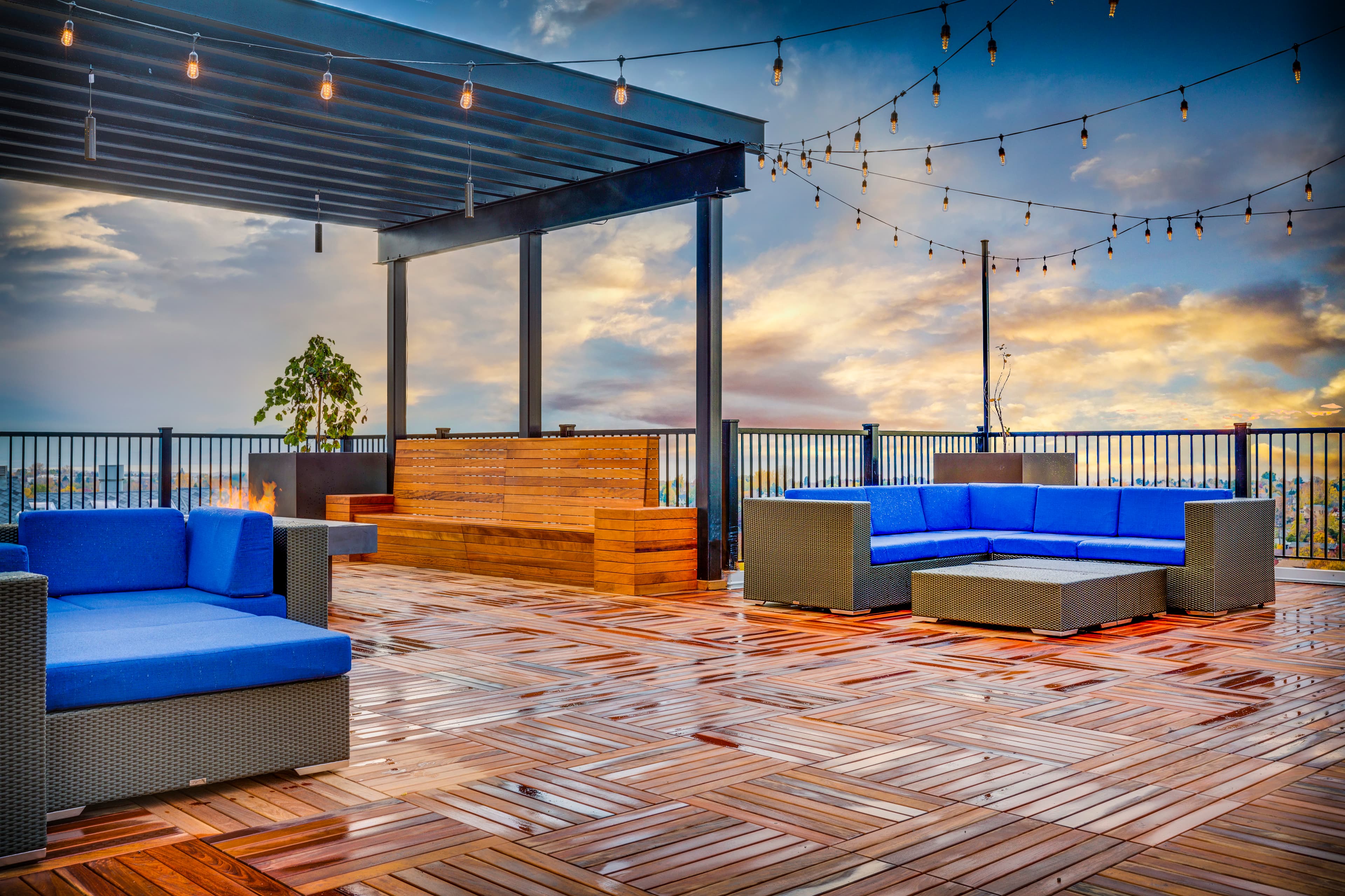 A rooftop patio at Midtown 360 Orem features blue cushioned wicker seating, wooden flooring, string lights overhead, and a wooden pergola with a dramatic sunset sky in the background.