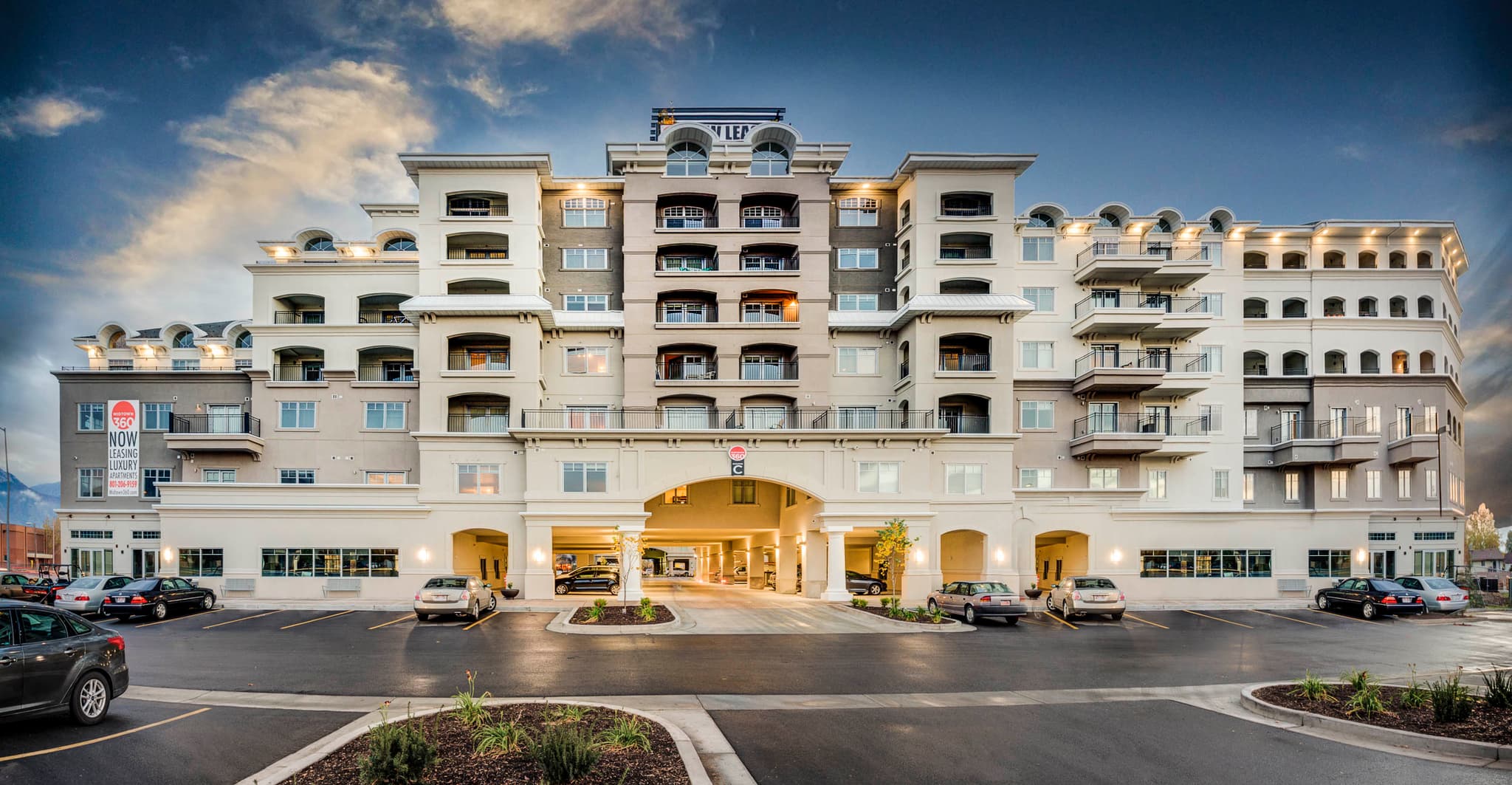 A modern, multi-story apartment building—Midtown 360 Orem—with balconies and arched entrances, surrounded by parked cars and landscaped greenery, stands under a partly cloudy sky at sunset.