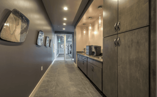 A modern, narrow kitchenette in the Moda Bonneville, Salt Lake City, featuring pendant lights, dark cabinets, a coffee machine, and decorative wall art. The hallway leads to a glass door with natural light at the far end.