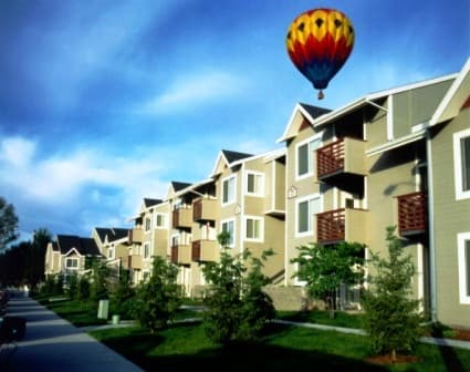 A colorful hot air balloon floats in the blue sky above Morrison Park Apartments Boise, where modern three-story tan buildings with balconies are lined by young trees and a welcoming sidewalk.