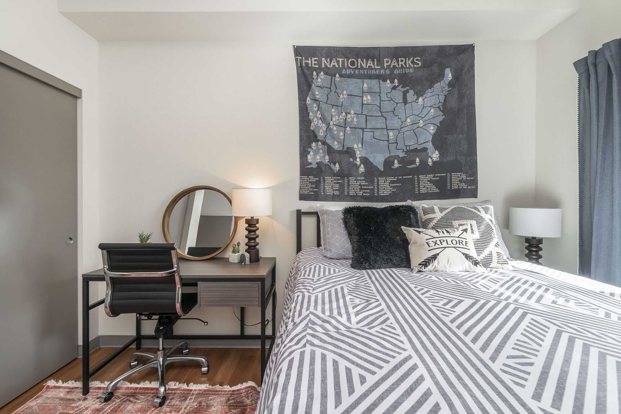 A modern bedroom at Patterson Social Eugene features a bed with geometric-patterned bedding, a map of national parks above, a small desk with a round mirror and lamp, and a black desk chair on a rug.