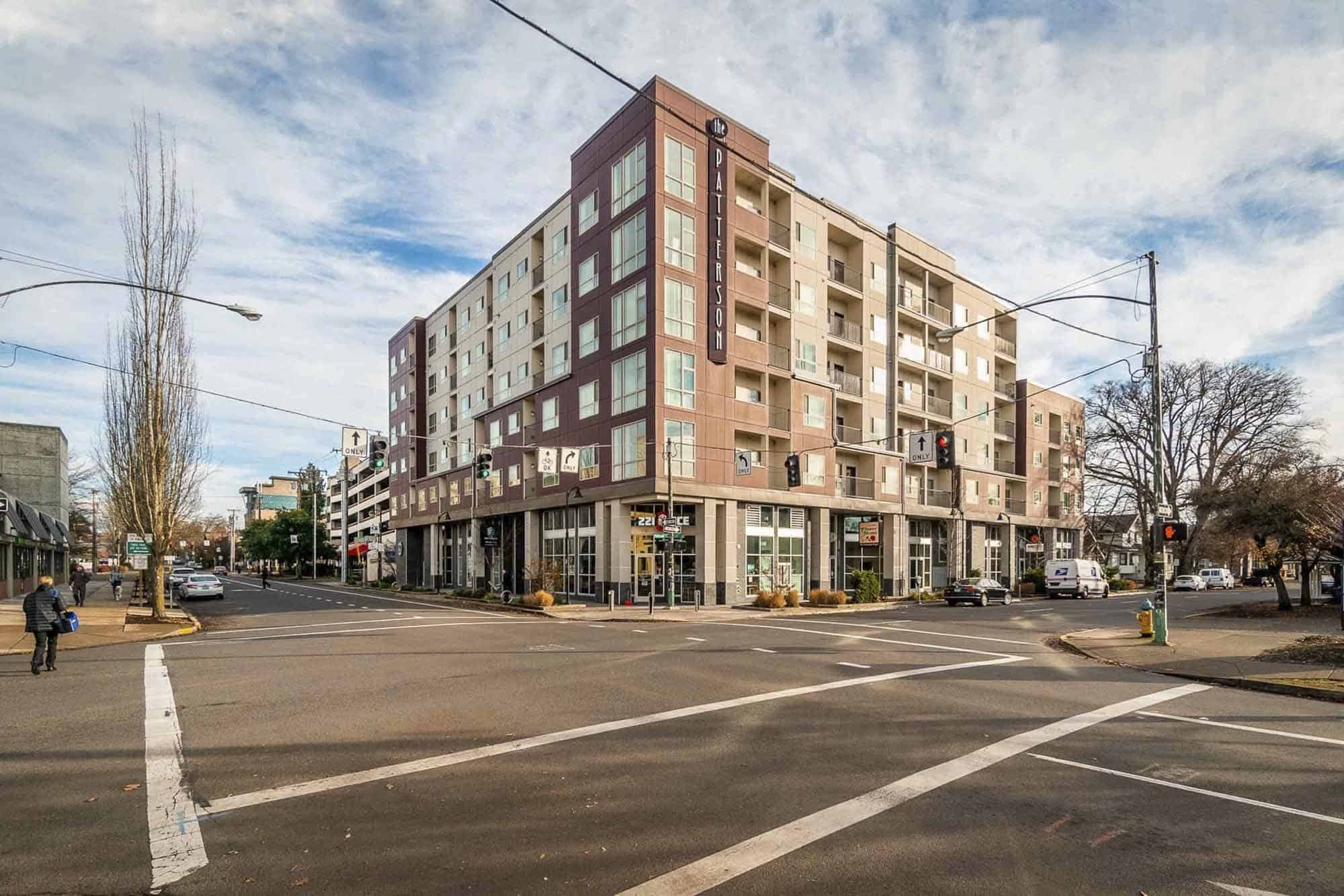 A modern, multi-story apartment building—Patterson Social Eugene—with ground-floor retail sits at a city street intersection on a partly cloudy day. Cars are parked along the street, and a few pedestrians are visible on the sidewalks.