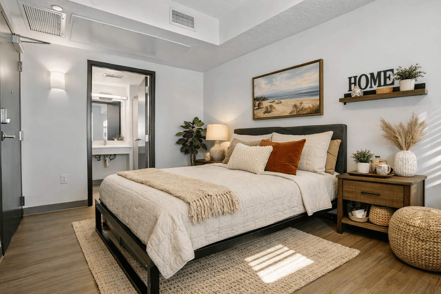 A cozy bedroom in Slopeside Village features a neatly made bed with beige bedding, accent pillows, a wooden nightstand, potted plants, and wall art above the bed. A bathroom is visible in the background, perfect for your Park City getaway.