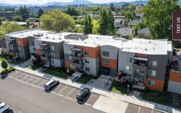 Aerial view of The Quad Corvallis, a modern three-story apartment building with gray and orange accents, surrounded by trees, residential houses, and a parking lot with cars in front.
