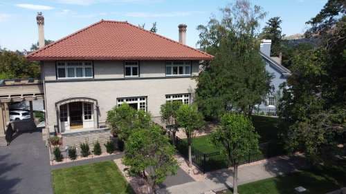 A two-story Renaissance House in Utah with a red tile roof, beige brick exterior, and large windows, surrounded by trees and a neatly manicured lawn, with a driveway and sidewalk visible in front.