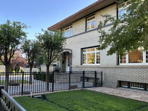 Two-story Renaissance House in Utah with large windows, surrounded by trees and a black metal fence. The neatly trimmed lawn and small walkway lead to the front entrance under a clear blue sky.