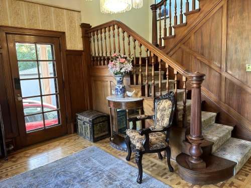 A vintage-style foyer in this Utah Renaissance House features a wooden staircase, ornate railing, patterned armchair, round table with a vase of flowers, decorative trunk, and a glass-paneled door letting in natural light.