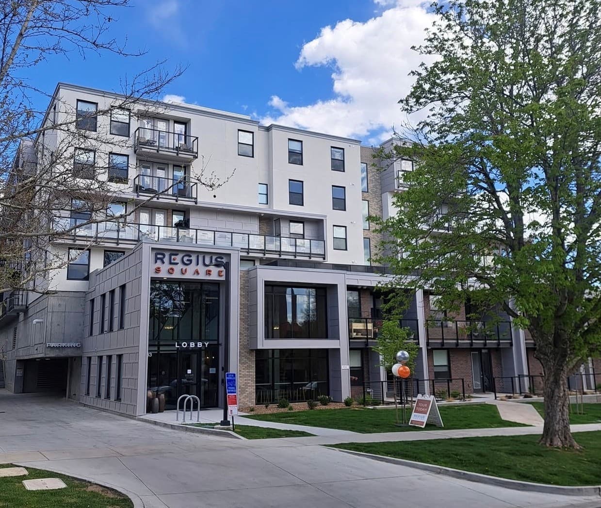 Regius Square is a modern multi-story apartment building featuring large windows, balconies, and a prominent lobby entrance. Lush trees, green grass, and a wide sidewalk fill the foreground beneath a blue sky with clouds.