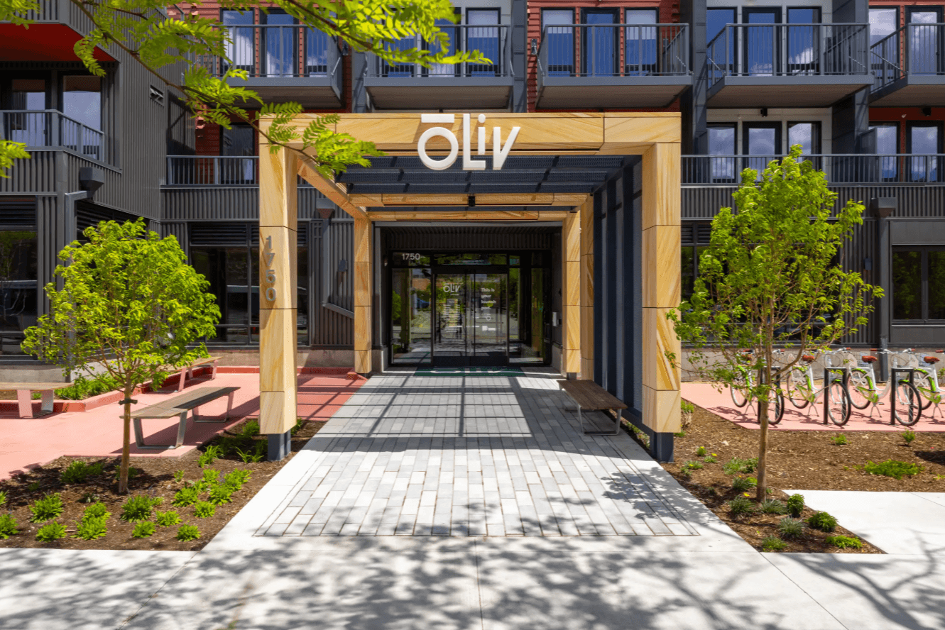 A modern apartment building entrance with a wooden pergola labeled Olly, surrounded by young green trees, benches, and bike racks, welcomes you to ōLiv Boulder Boulder through sleek glass doors.
