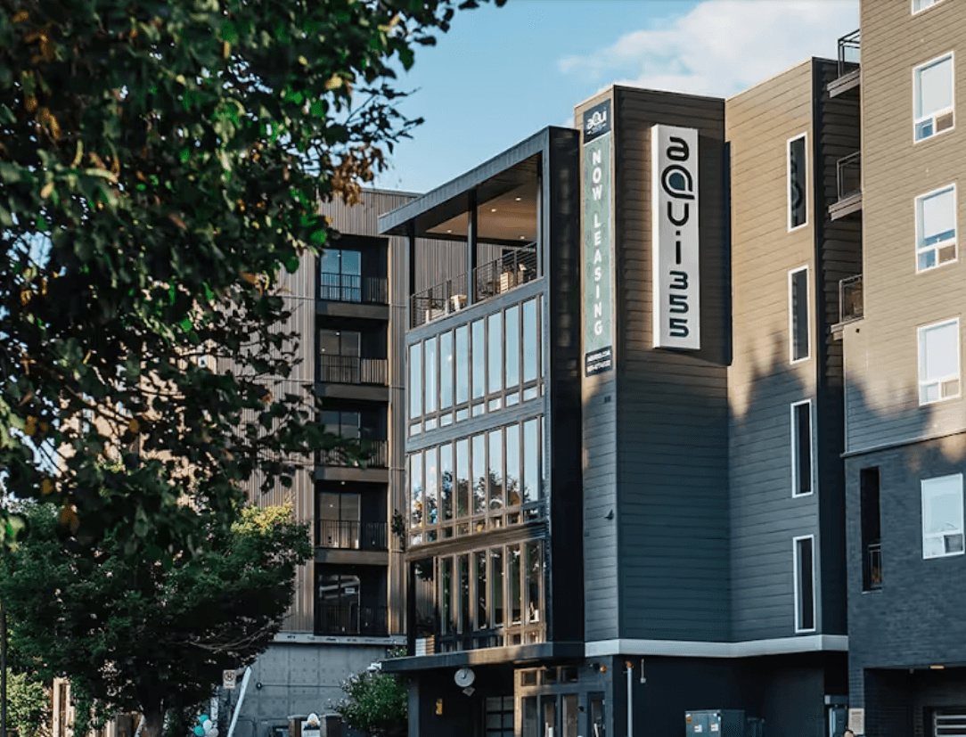 Modern apartment building with large windows and balconies, surrounded by trees. A vertical sign on the building reads AQUI 35 and a banner says Now Leasing. The sky is partly cloudy.