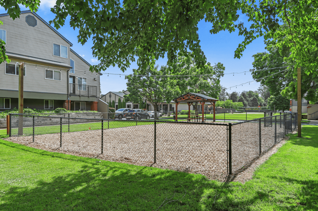 A fenced-in area with wood chips, string lights overhead, and a wooden gazebo sits on the grassy lawn at Ducks Village near an apartment building and parked cars under sunny skies.