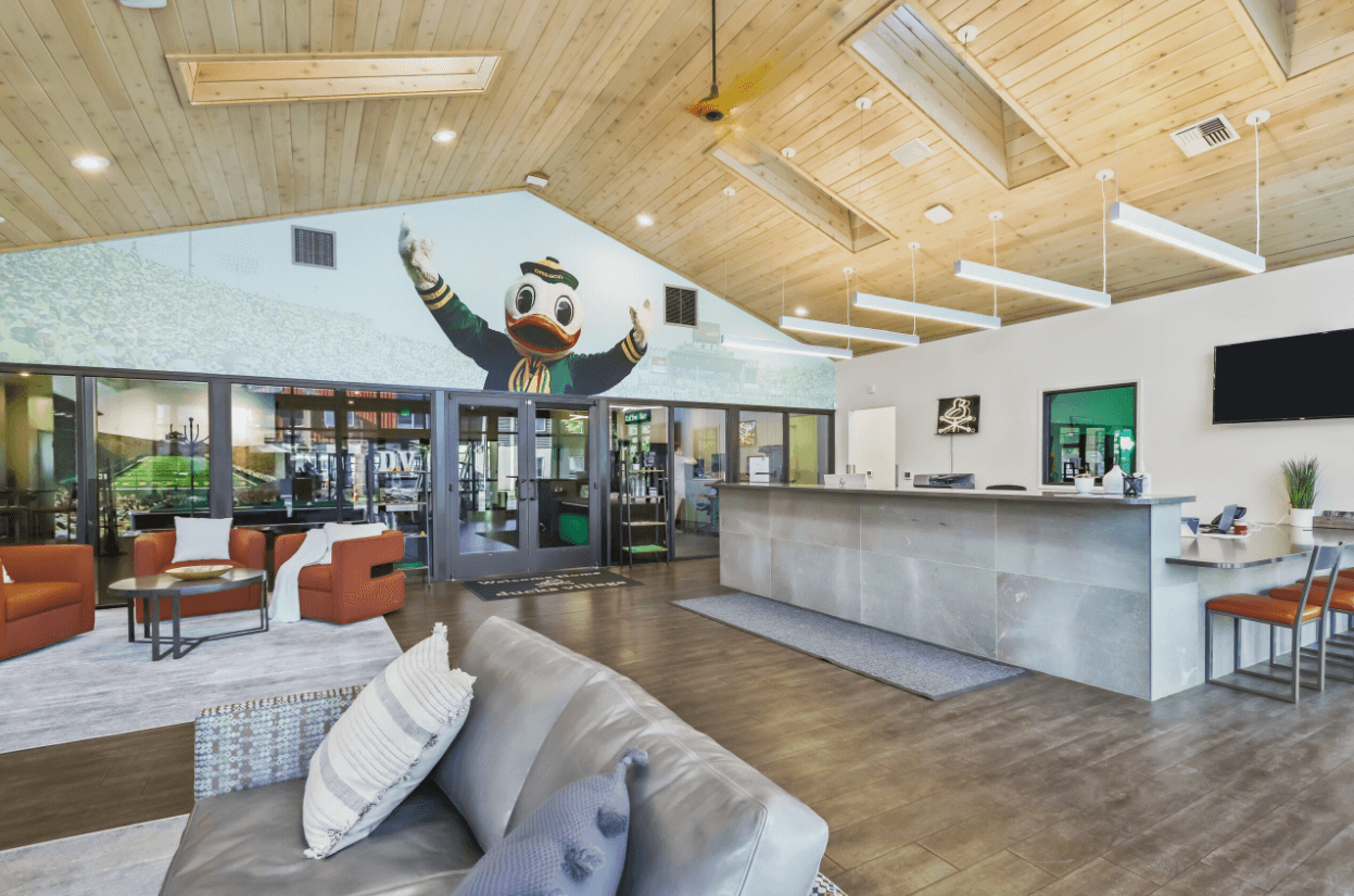 Modern lobby at Ducks Village with wood ceiling, gray tile floor, sleek reception desk, lounge seating, and a mural of a waving duck mascot above glass doors. Bright lighting and contemporary decor create a welcoming atmosphere.