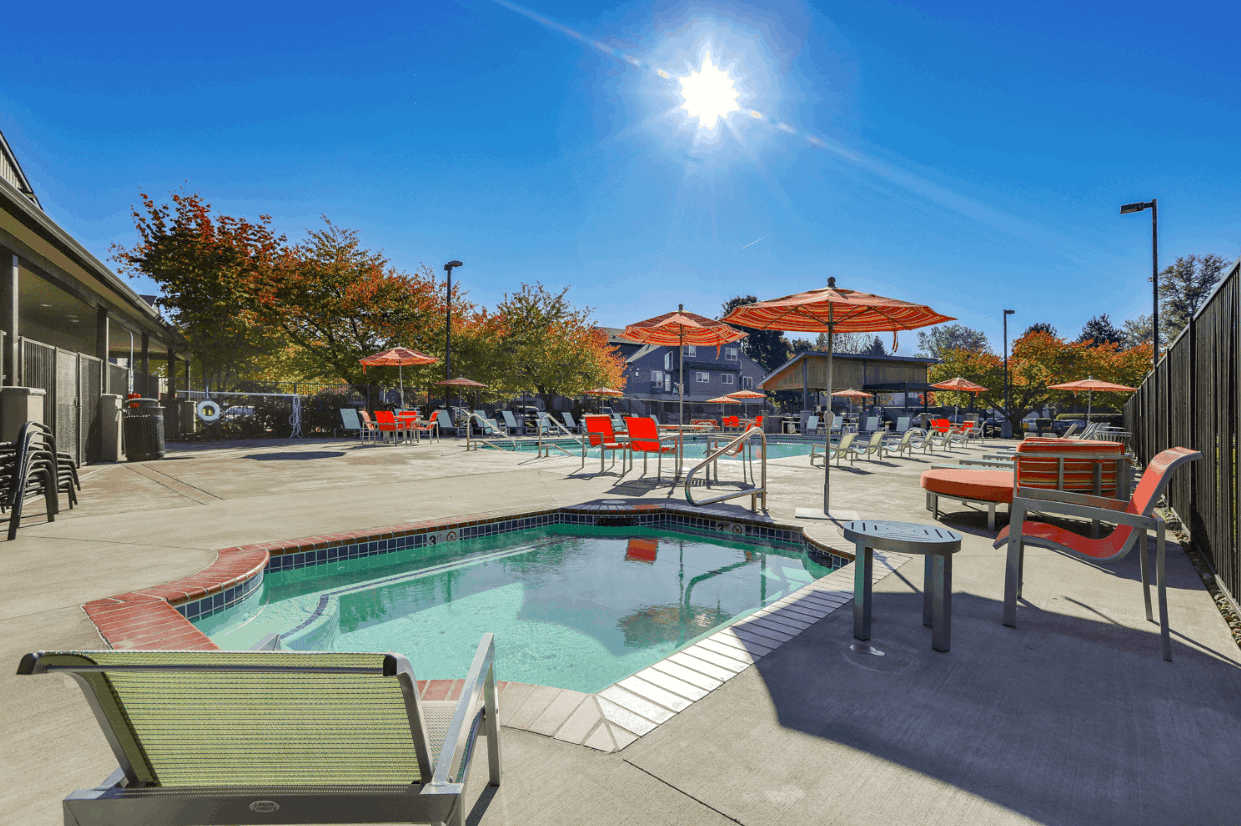 Ducks Village outdoor pool area on a sunny day with a small hot tub in the foreground, red chairs and orange umbrellas scattered around, trees and buildings in the background, and bright blue sky overhead.