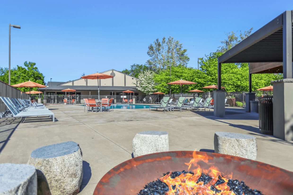 Outdoor pool area at Ducks Village with lounge chairs, orange umbrellas, and a fire pit in the foreground. A shaded seating area and trees surround the pool under a clear blue sky.