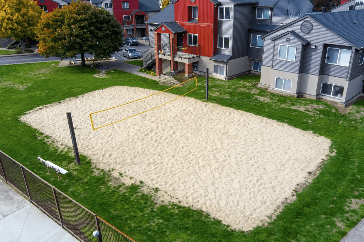 A sand volleyball court with a yellow net is surrounded by grass and located next to Ducks Village’s multi-story apartment buildings. Trees and parked cars are visible nearby.
