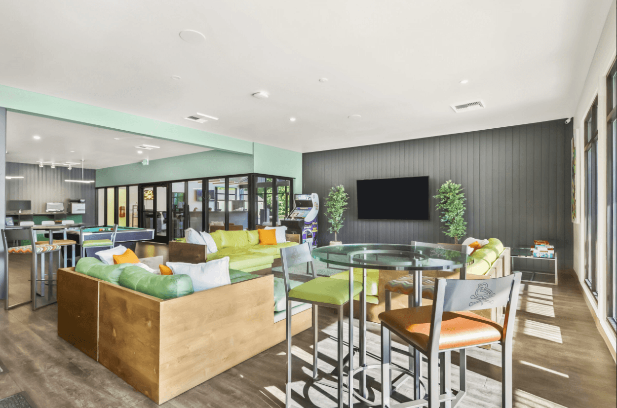 A modern lounge at Ducks Village with colorful seating, glass tables, a large wall-mounted TV, arcade game machine, and potted plants. Sunlight pours in through large windows, and there are games stacked in the corner.