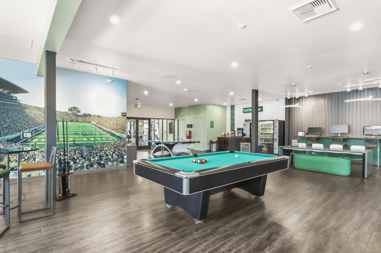 A modern game room at Ducks Village features a pool table at the center, a mural of a football stadium, computers on desks, green accents, wood flooring, and large windows that let in plenty of natural light.