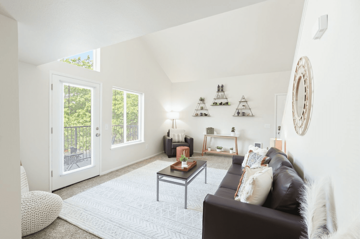 Bright, modern living room in Ducks Village with white walls, a dark sofa, a light rug, and a coffee table. Large windows and a glass door open to a balcony with lush greenery. Minimalist decor and wall shelves enhance the airy feel.