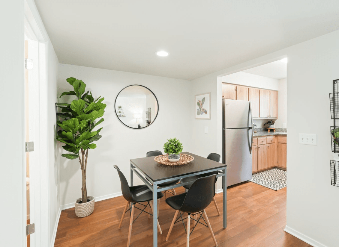 A modern dining area at Ducks Village features a black table, four black chairs, a round mirror on the wall, a large potted plant, and a view into a kitchen with wooden cabinets and stainless steel appliances.