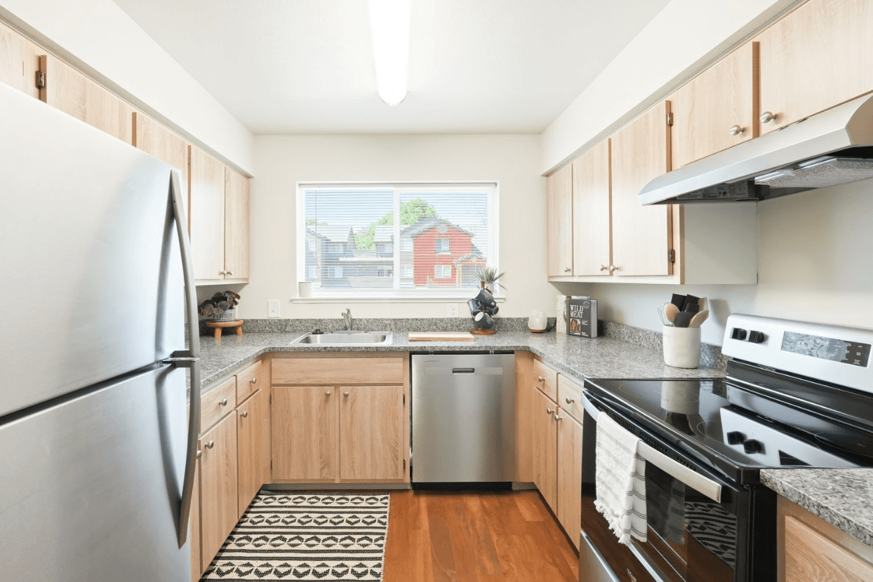 Modern kitchen at Ducks Village with light wood cabinets, stainless steel appliances, granite countertops, a patterned rug, and a large window letting in natural light.