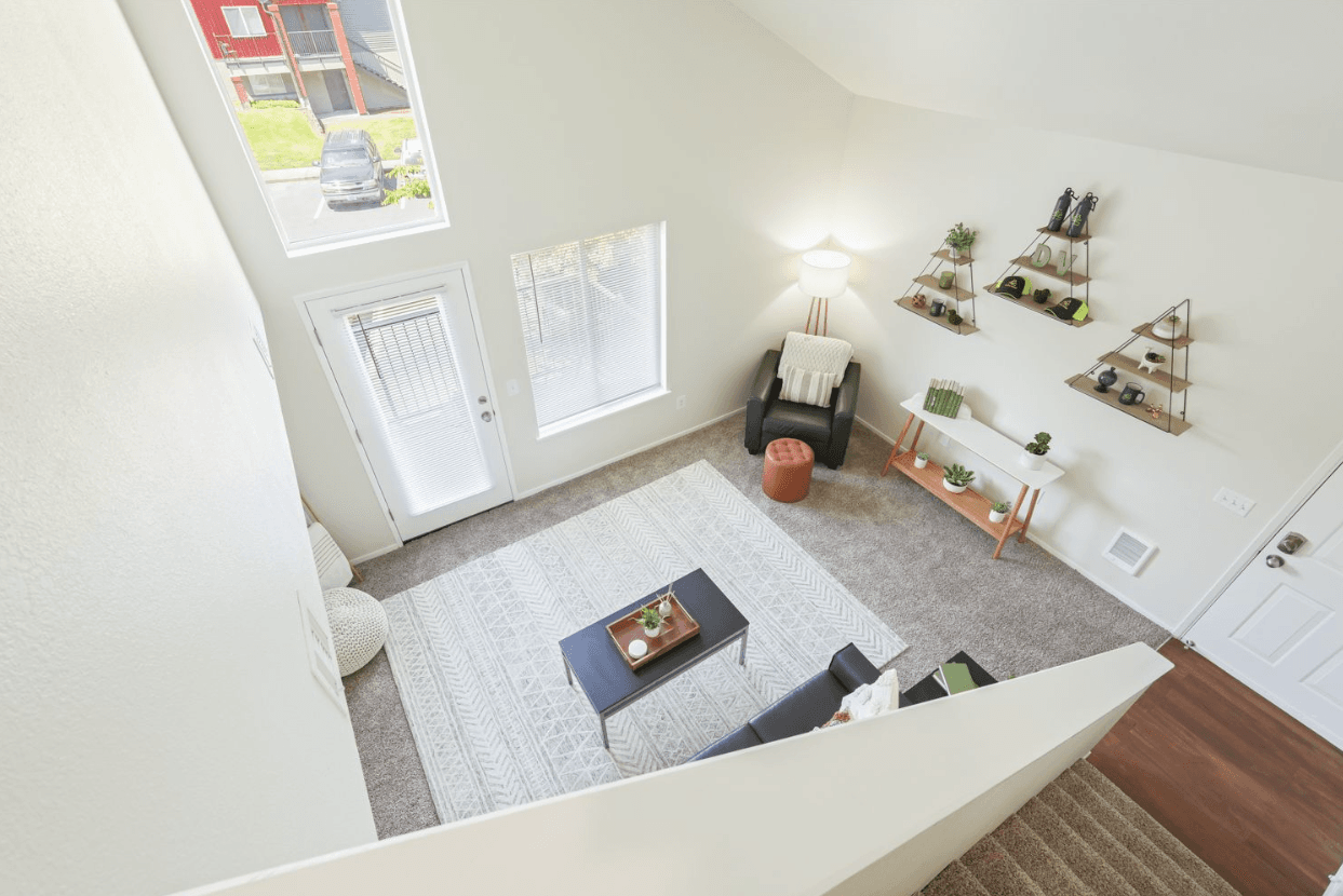 Bright living room seen from above at Ducks Village, with large windows, a white rug, gray couch, coffee table, armchair, floor lamp, wall shelves with plants, and a door leading outside. Carpeted floor with wood near the entrance.