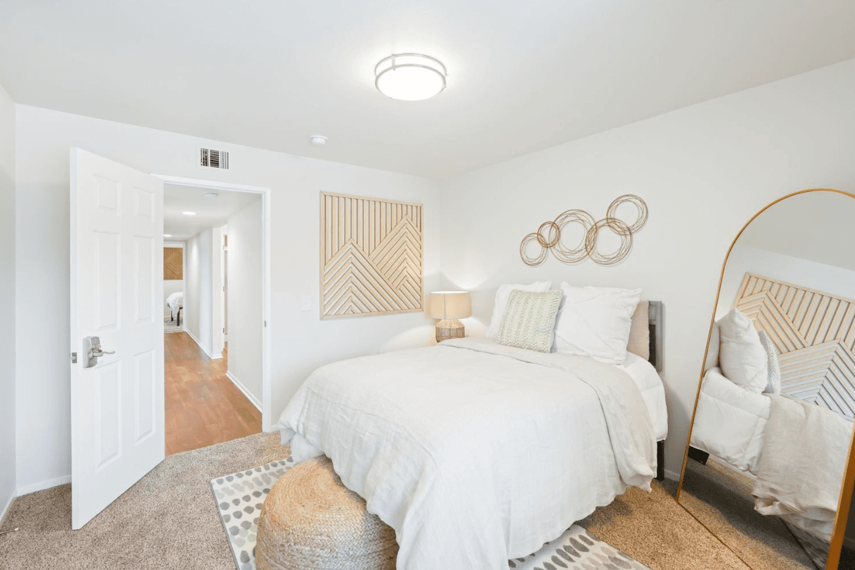 A bright, minimalist bedroom in Ducks Village features a neatly made bed, neutral bedding, round gold mirror, woven pouf, bedside lamp, and geometric wall art. The open door reveals a hallway with wood flooring.