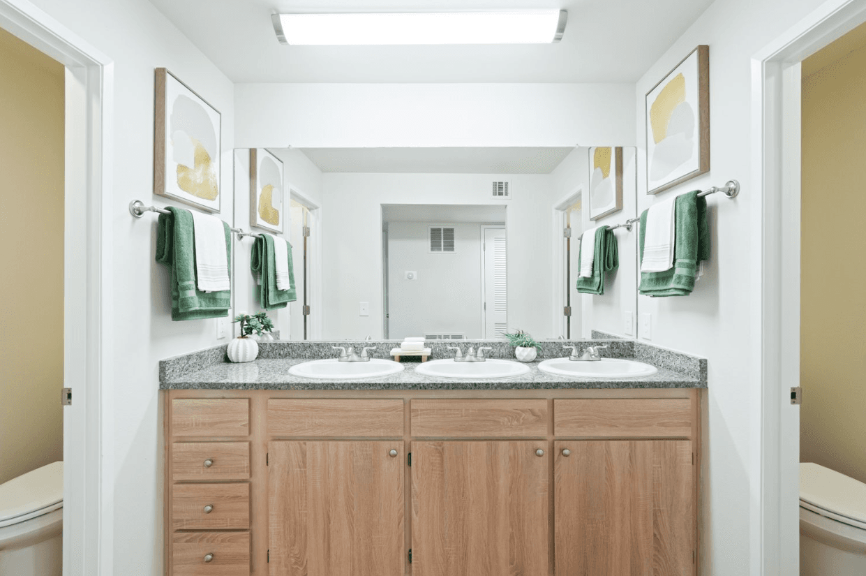 A modern bathroom at Ducks Village features a double sink vanity, wood cabinets, and a large mirror. Each sink has green and white towels, minimal decor, and framed abstract art above. Two doorways are visible on either side.