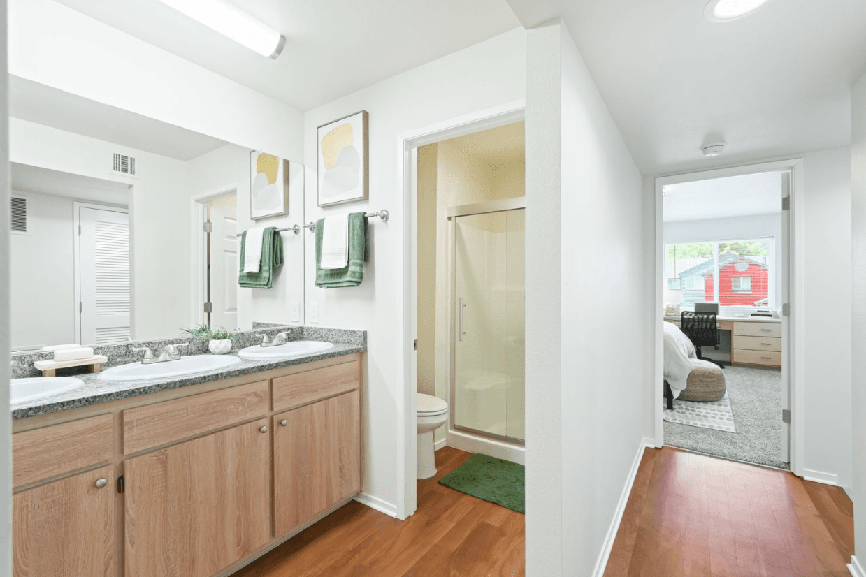 Bright bathroom at Ducks Village with a double sink vanity, large mirror, and green towels. A shower with glass doors is next to the toilet. Wood flooring leads to a bedroom with a bed, desk, and window.