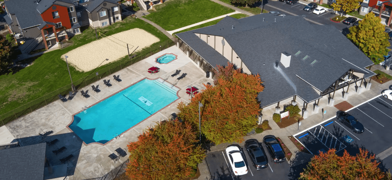 Aerial view of Ducks Village apartment complex with a swimming pool, hot tub, lounge chairs, sand volleyball court, parking lot, and autumn trees. Nearby buildings and cars are also visible.