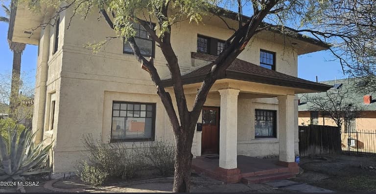 Charming two-story beige stucco house at 833 N 4th Ave features dark trim, a covered front porch with square columns, and a large tree in the yard. Some shrubs and an agave plant are visible near the entrance.