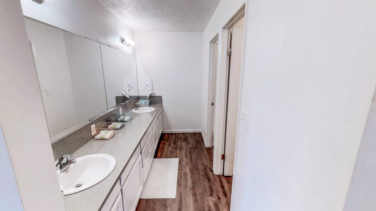 Bright bathroom at Somerset Apartments Rexburg with double sinks, a large wall mirror, white countertops, wooden floor, and white walls. Two doorways are on the right and decorative items sit near the sinks.
