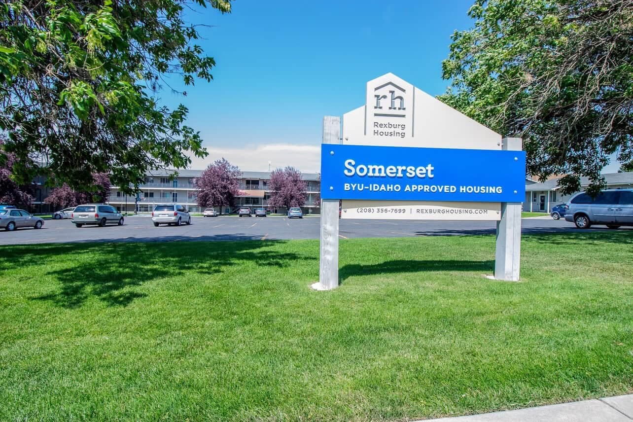 A sign on a grassy lawn reads “Somerset BYU-Idaho Approved Housing” with a Rexburg Housing logo. Somerset Apartments Rexburg and a parking lot are visible in the background under a clear sky.