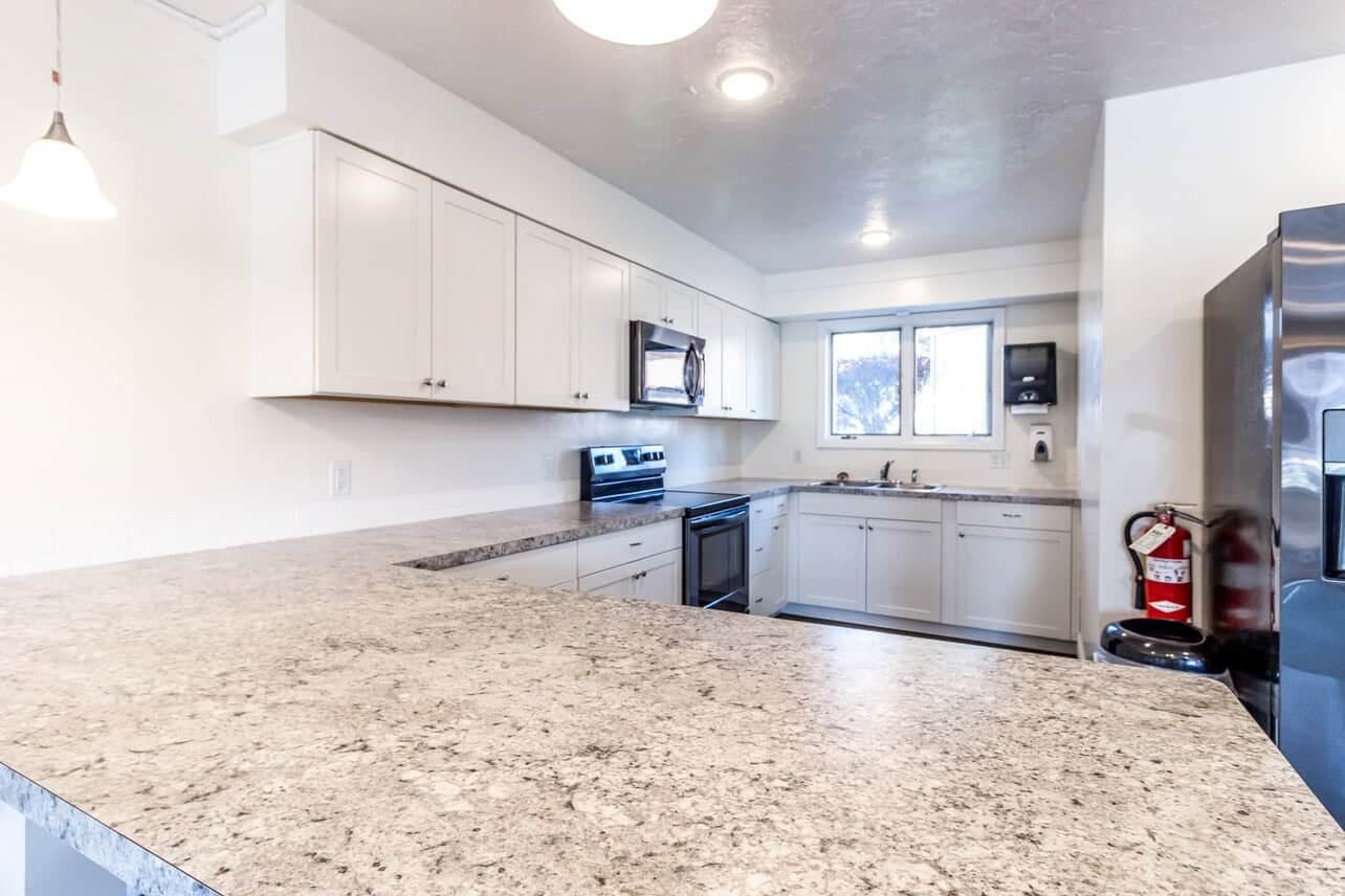 Modern kitchen at Somerset Apartments Rexburg with white cabinets, stainless steel appliances, a granite countertop, window above the sink, and a black refrigerator. A fire extinguisher is mounted near the refrigerator for safety.